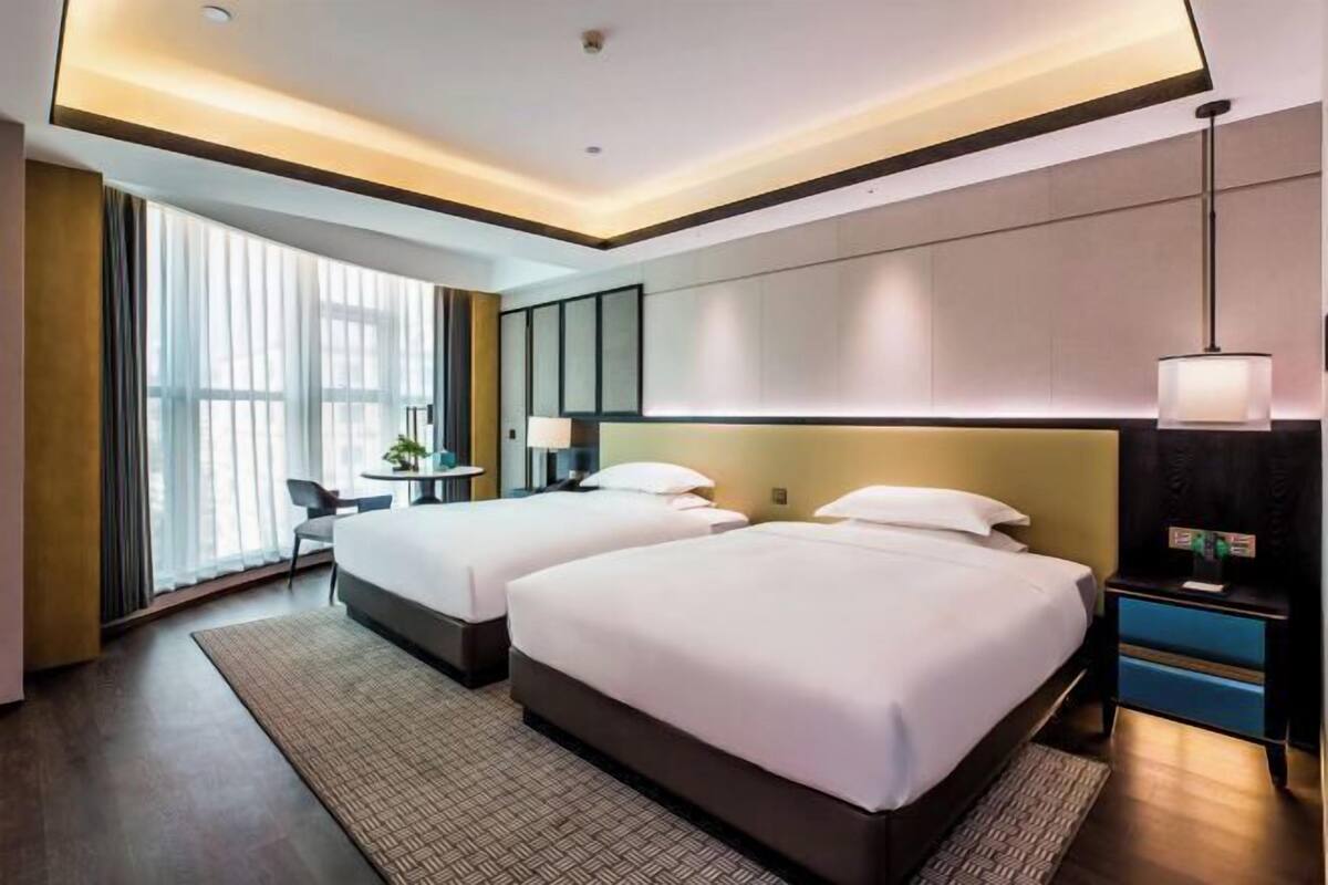 A hotel room with two beds, a desk, a chair, and a large window with curtains.