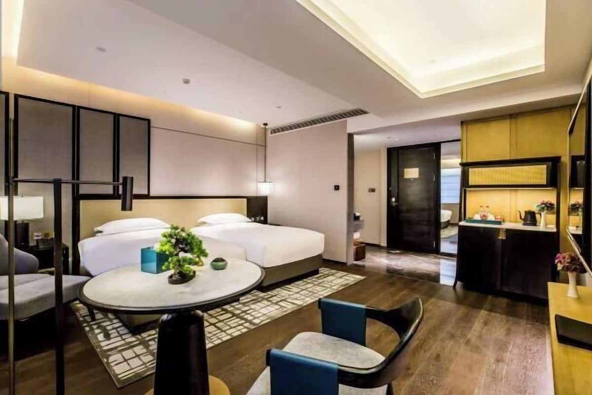 A modern hotel room with a large bed, a round dining table, a sofa, and a small kitchen area.
