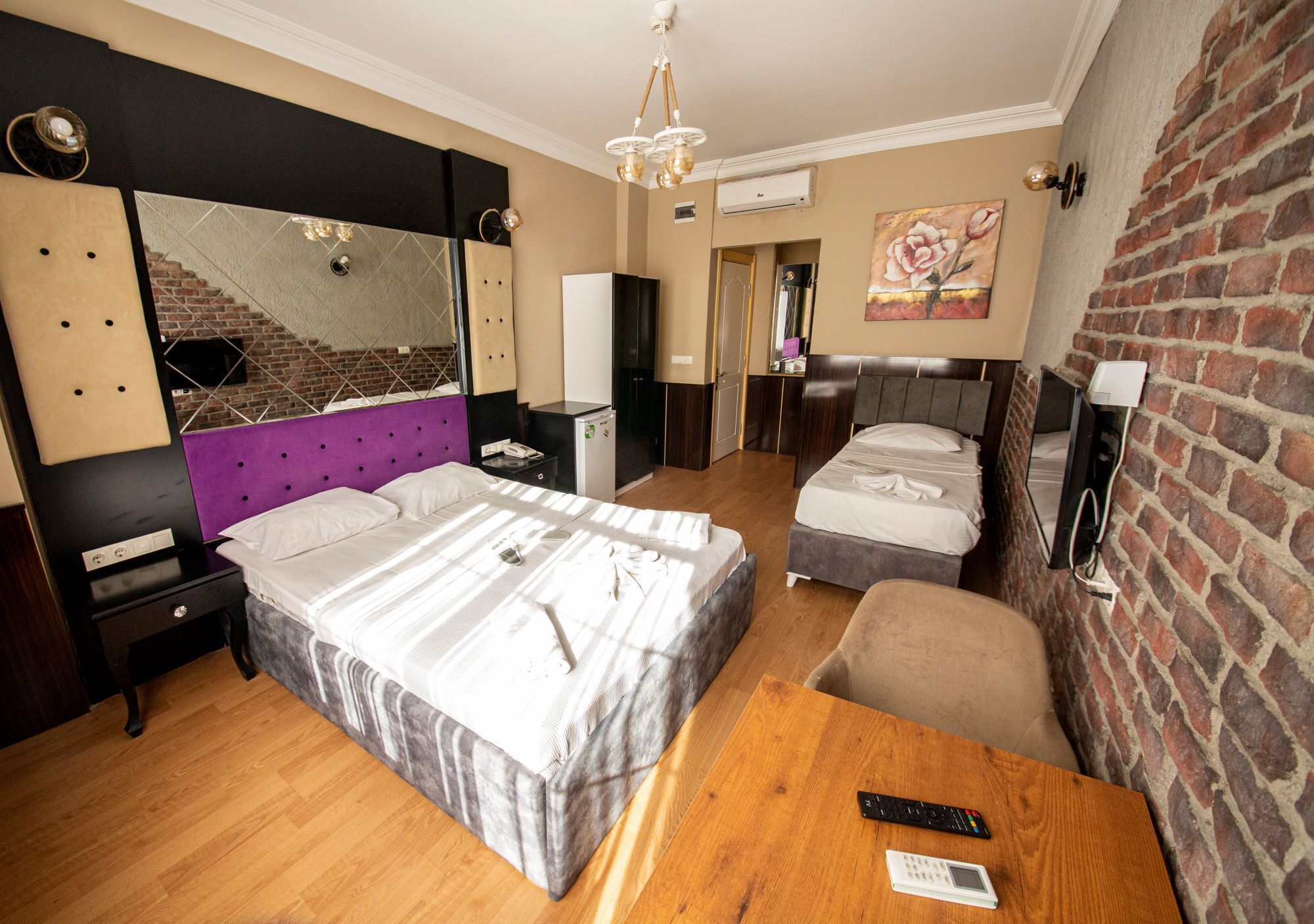 A hotel room with a large bed, a smaller bed, a wooden table, a chair, a mirror, a TV, and a brick wall.