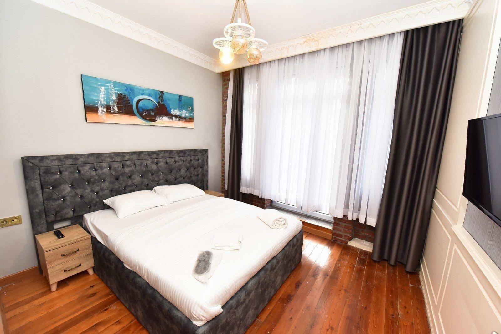 A bedroom with a large bed, a nightstand, a TV, a painting, and a window with curtains.