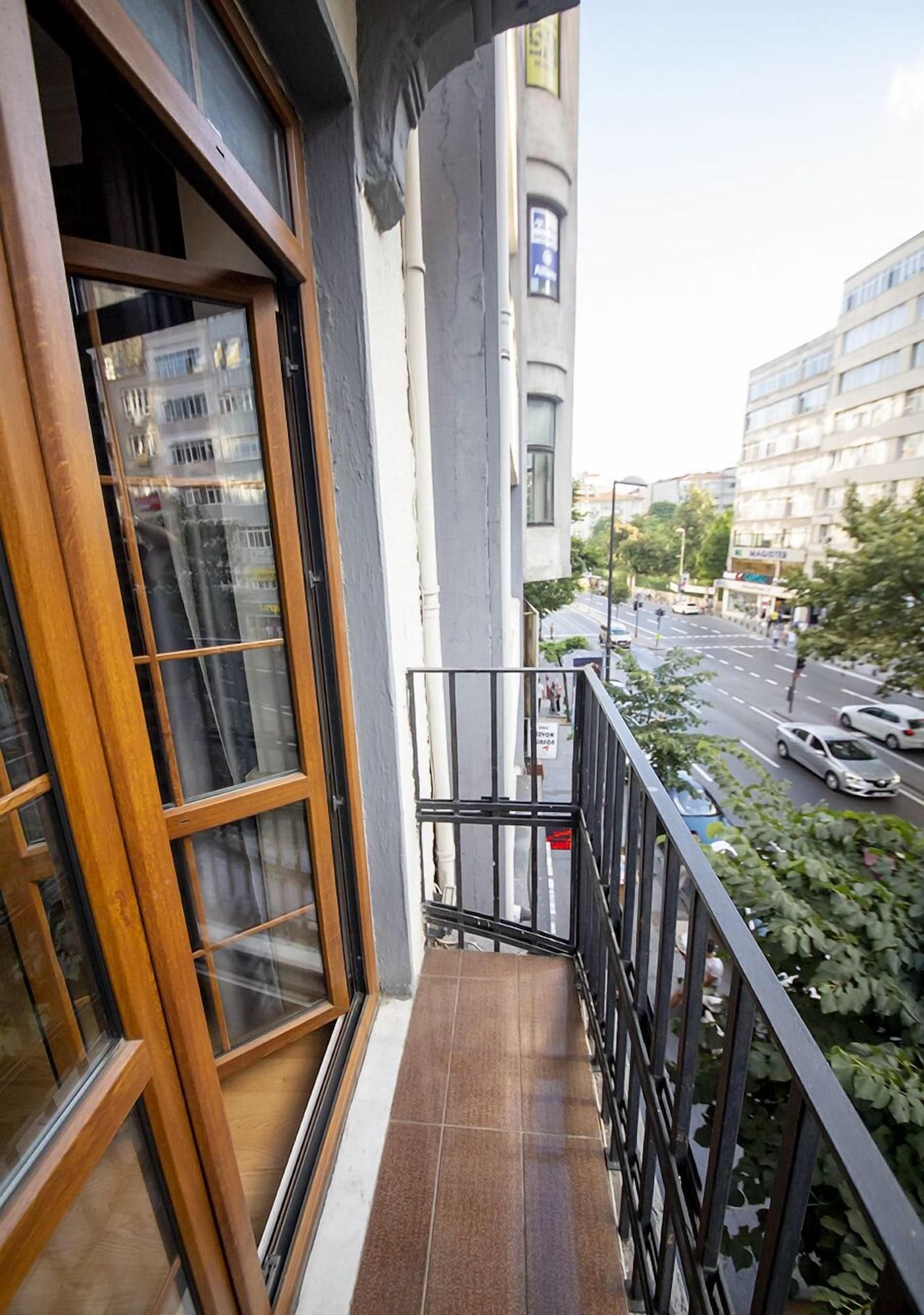 A balcony with a wooden-framed window, a metal railing, and a view of a city street with buildings and cars.