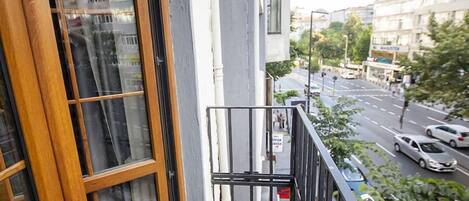 A balcony with a wooden-framed window, a metal railing, and a view of a city street with buildings and cars.