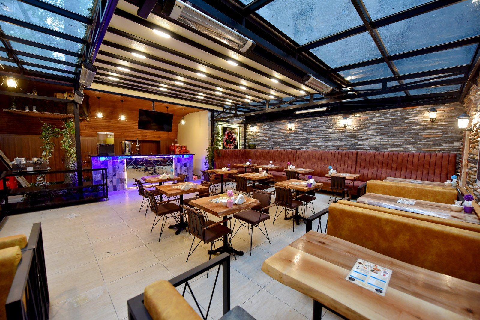 A restaurant with a glass ceiling, wooden tables and benches, and a stone wall.