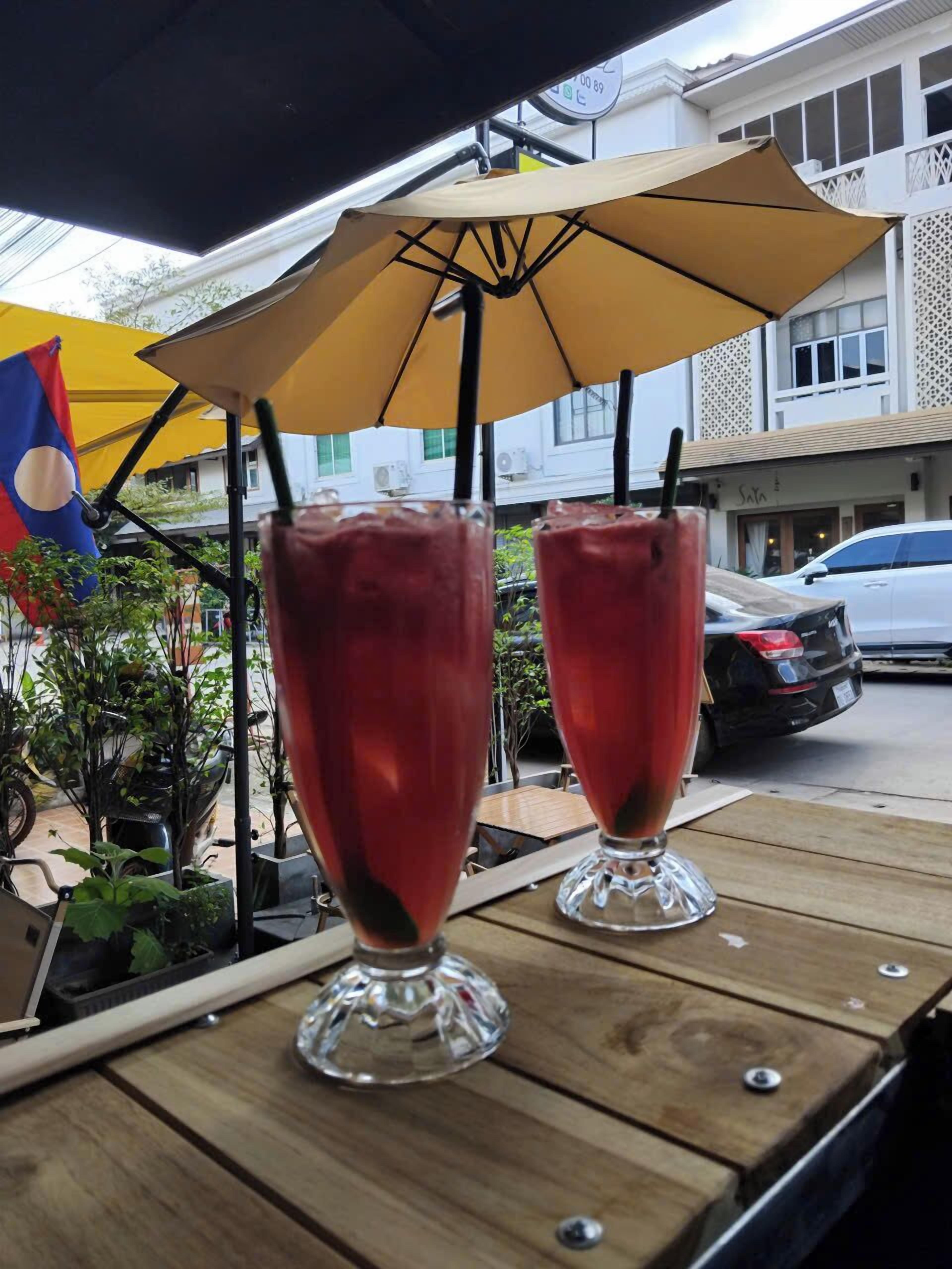 Deux grands verres d’une boisson rouge sont posés sur une table à l’extérieur, à l’ombre d’un parasol jaune.