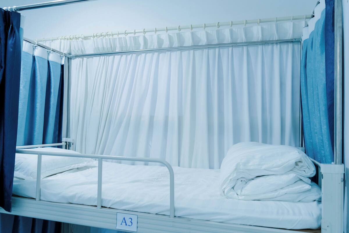 Ein Krankenzimmer mit einem Einzelbett mit der Aufschrift A3, weißen Vorhängen und einem Metallgestell.