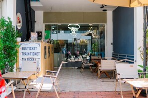 An outdoor seating area with tables and chairs, a sign for bus, train, and air tickets, and a glass door entrance. - Happy Home Hostel (Vientiane)