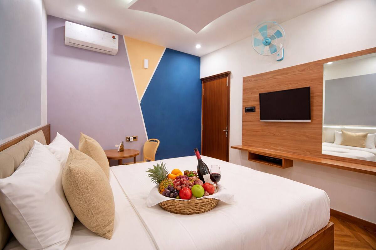 Een hotelkamer met een bed, een klein tafeltje met fruit en wijn, een aan de muur bevestigde televisie en een plafondventilator.