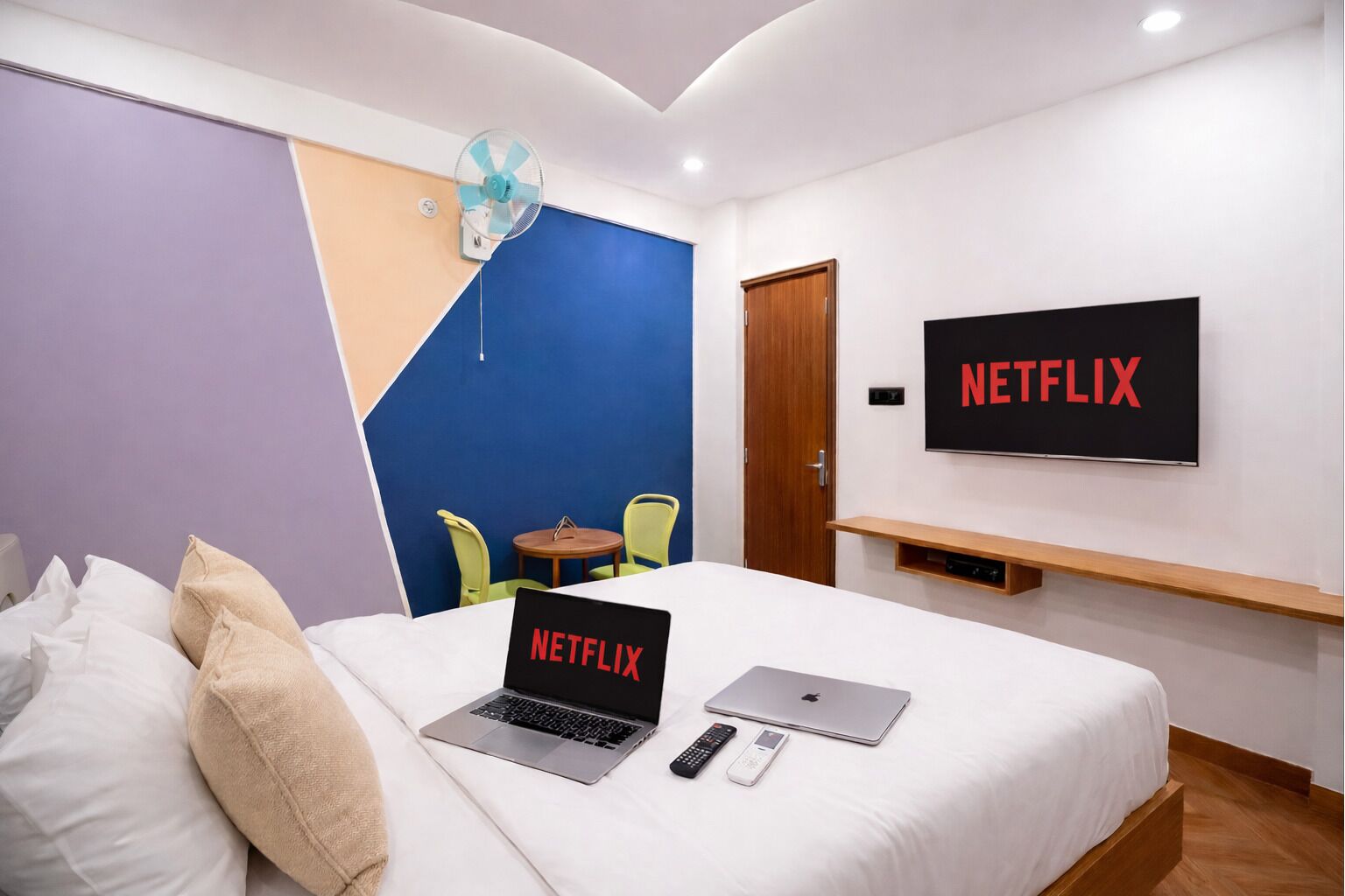 A hotel room with a bed, a laptop, a remote control, a phone, a wall-mounted TV with 'NETFLIX' on it, a fan, and a door.
