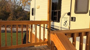 A covered deck area with a wooden railing, a white RV parked adjacent, and a clear view of the surrounding trees and sky. - Sunset Hideaway (Springtown)