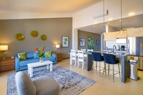 Un salon moderne avec un canapé bleu, une table basse et un coin repas comprenant des chaises blanches et un bar.