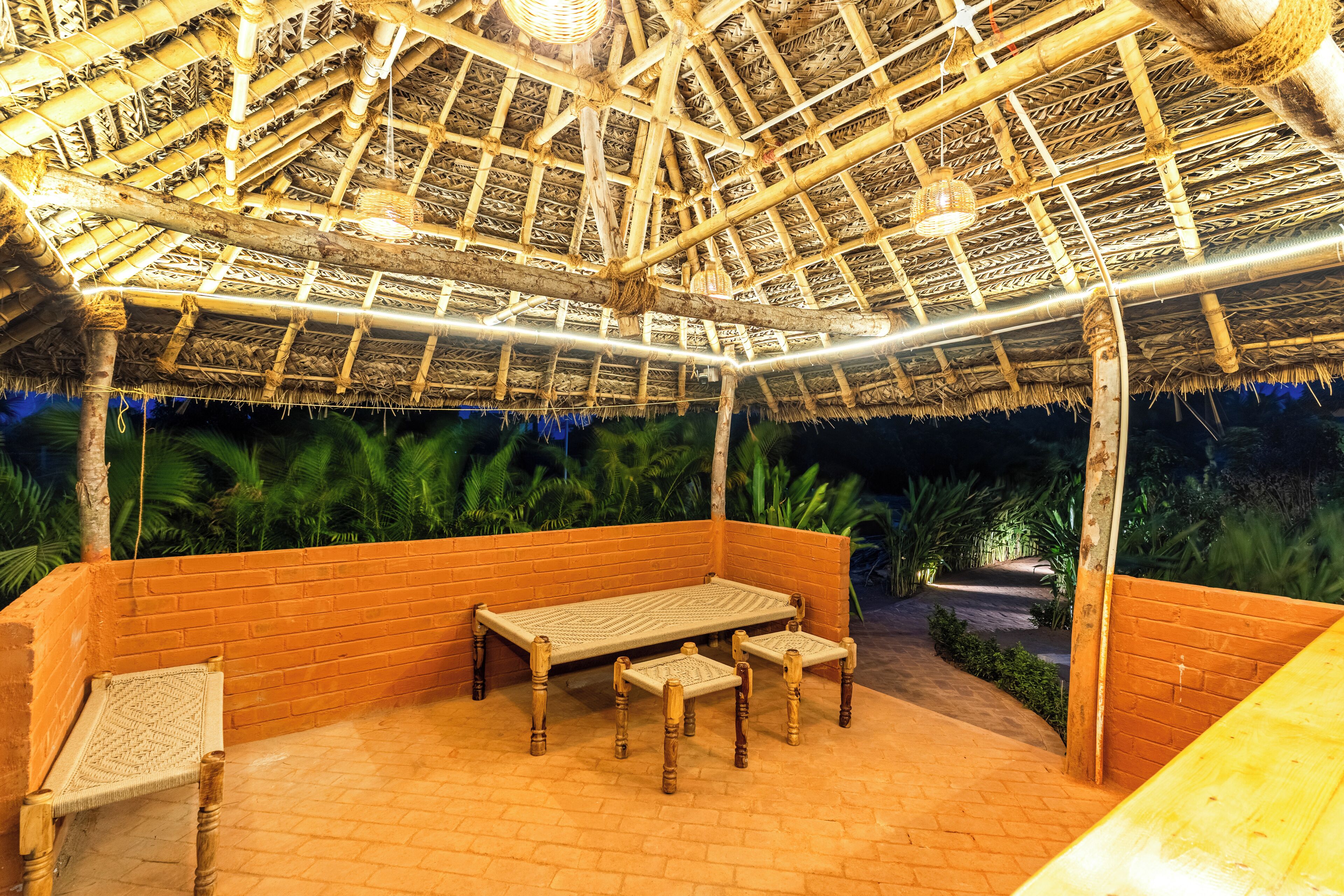 A covered outdoor seating area with a thatched roof, wooden furniture, and a brick wall.