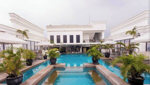 A pool area with a central pool, surrounded by potted plants, and a building with a balcony in the background.