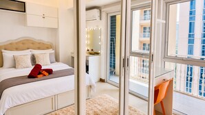 A bedroom with a bed, a desk, a mirror, and a view of the outside. - AdahkuMaya Luxury Suites Plus (Taguig)