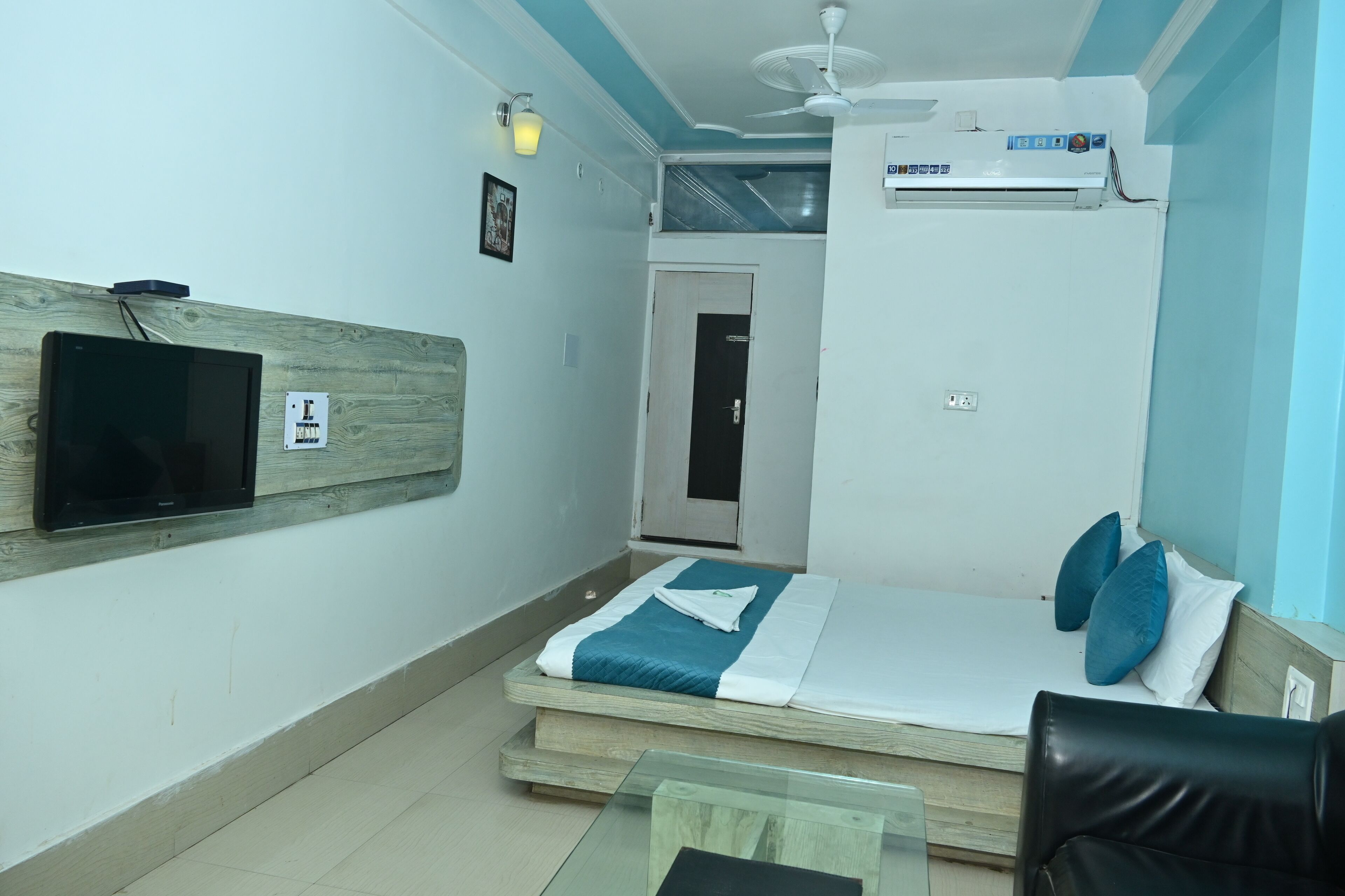 A hotel room with a bed, a TV mounted on the wall, a ceiling fan, and an air conditioning unit.