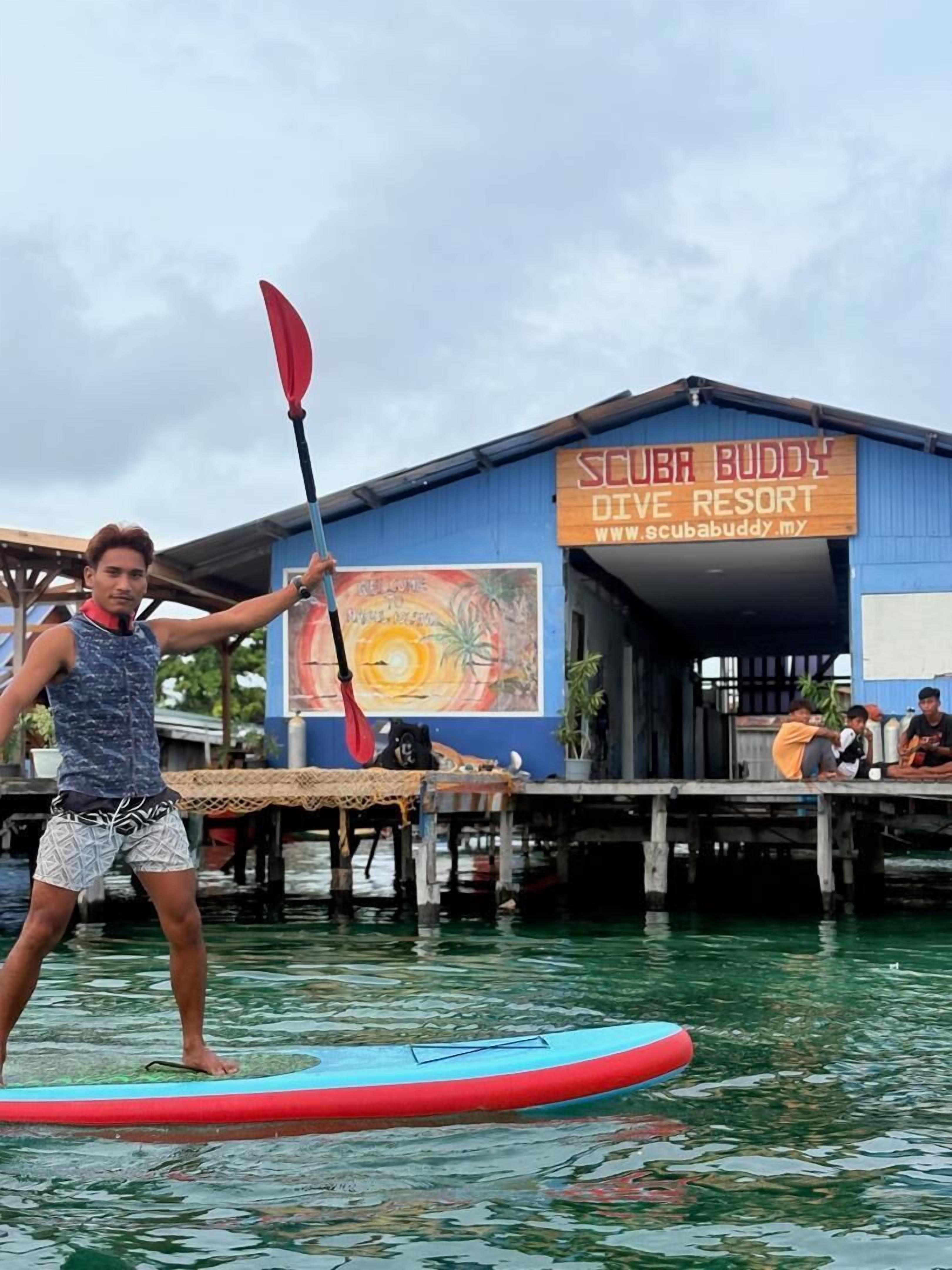 A person paddleboarding in front of a building labeled "SCUBA BUDDY DIVE RESORT".
