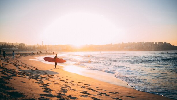 Una spiaggia con impronte che conducono all'acqua, una persona che tiene un’asse da surf e uno skyline cittadino sullo sfondo.