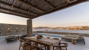 Ein Holztisch mit Stühlen vor einer Steinwand, mit Blick auf einen Pool und eine schöne Aussicht auf eine Küstenstadt.