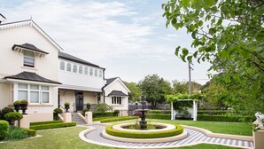 Uma casa de dois andares com um jardim bem cuidado, uma fonte e um caminho com padrão xadrez.