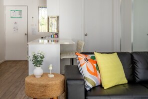 A living room with a black sofa, a round woven ottoman, a small white vase, and a colorful pillow.