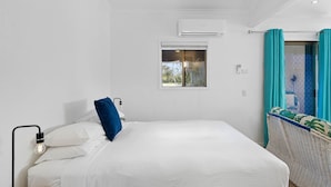 Ein Schlafzimmer mit einem Bett, einem Fenster, einer Klimaanlage, einer Nachttischlampe und einem Korbstuhl mit Blick ins Freie.
