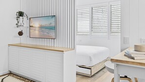 A modern bedroom with a bed, a bedside table with books, a wall-mounted TV, and a window with blinds. - Studio4: Bondi Chic Interiors with Rooftop Pool (Bondi Junction)