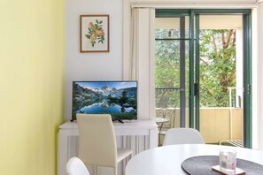 A room with a TV on a white stand, a dining table with chairs, and a balcony with a view of greenery. - Serene Studio with Balcony in Prime Location (Wollstonecraft)