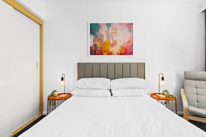 A modern bedroom with a large bed, two bedside tables with lamps, a grey upholstered chair, and a colorful abstract painting on the wall.