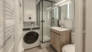 Casa de banho compacta com máquina de lavar roupa, cabine de duche em vidro e toucador de madeira.