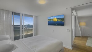 Habitación de hotel con una cama grande, un televisor en la pared, un balcón con vistas al mar y una ventana con cortinas.