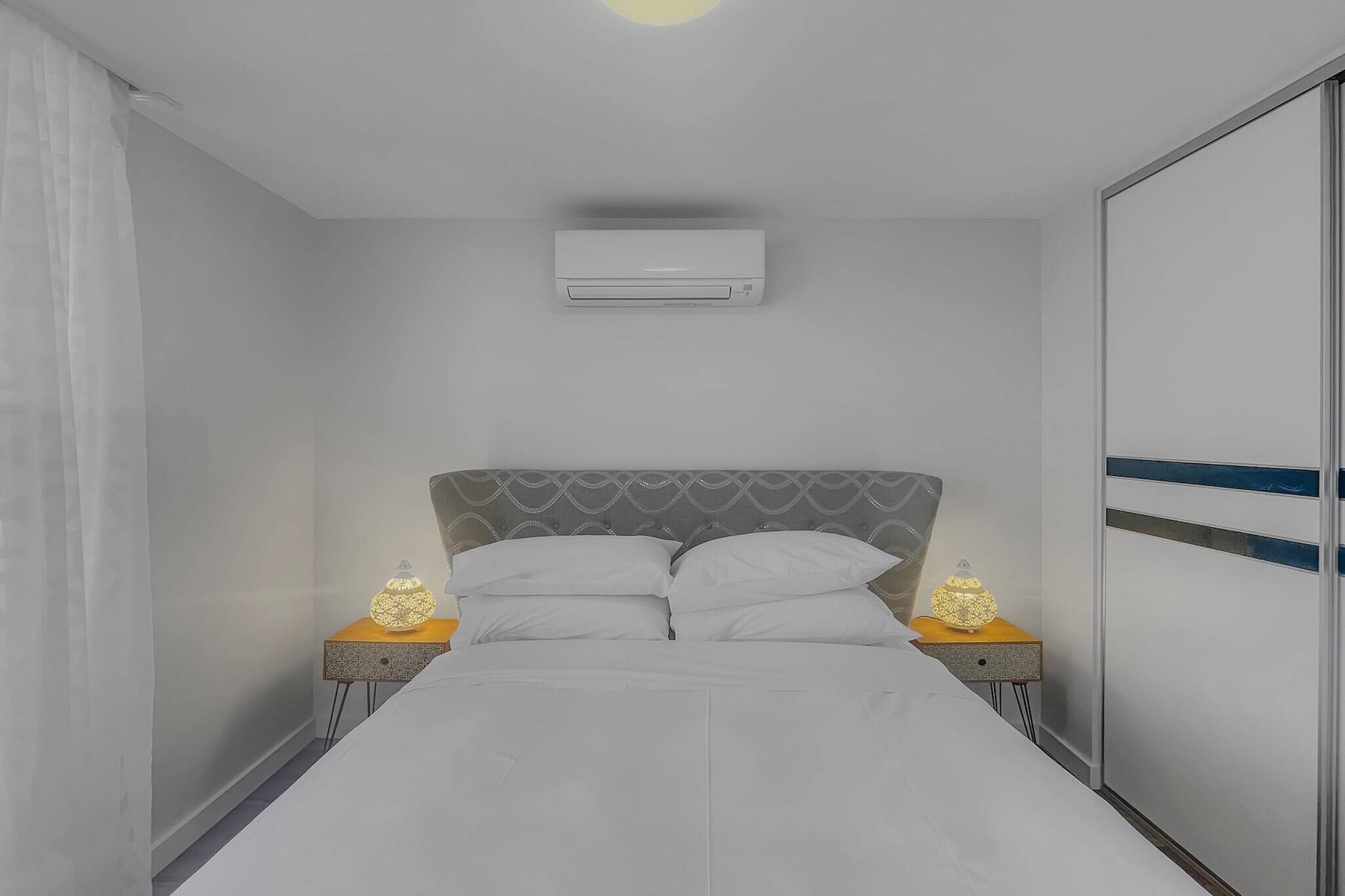 A bedroom with a bed, two bedside tables with lamps, a ceiling air conditioner, and a wardrobe.