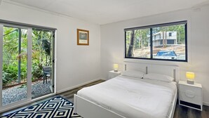 Ein Schlafzimmer mit einem Bett, zwei Nachttischen mit Lampen, einem Fenster mit Blick auf Grünflächen und ein Gebäude sowie eine Tür zu einem Außenbereich.