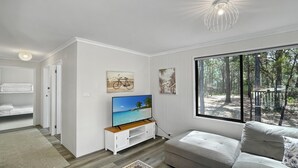 Ein modernes Wohnzimmer mit Fernseher, einer Couch, einem Fenster mit Blick auf einen Wald und einem Etagenbett im Hintergrund.