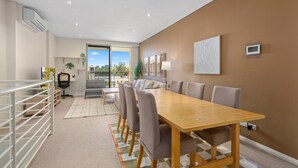 A modern dining area with a long wooden table, cushioned chairs, and a view of the city through large windows. - Silverwater Stay Near Parks and Cafés (Silverwater)