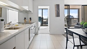 A modern kitchen with a large island, a dining area with a table and chairs, and a view of the city through large windows. - Luxury 2-Bed Apartment in Melbourne's Heart (Melbourne)