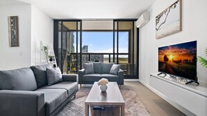 A modern living room with a grey sofa, a coffee table with a vase, a flat-screen TV on a stand, and a large window with a city view. - Luxury 2-Bed Apartment in Melbourne's Heart (Melbourne)