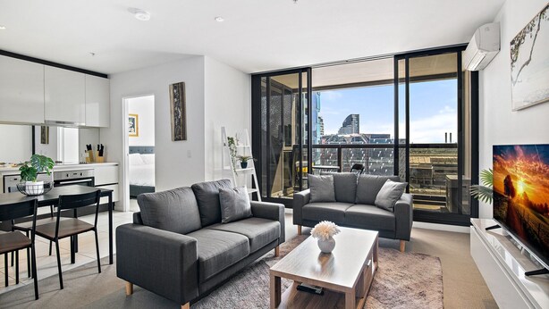 A modern living room with two grey sofas, a coffee table, and a TV. There is a kitchen area with white cabinets and a dining table. Large windows offer a city view. - Luxury 2-Bed Apartment in Melbourne's Heart (Melbourne)