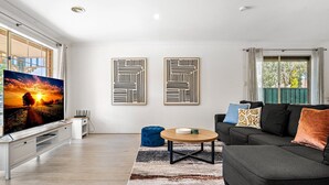Una sala de estar moderna con un televisor de pantalla plana, un sofá negro y una mesa de centro. Se observan dos obras de arte abstracto en la pared y una ventana con cortinas.