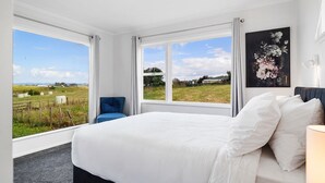 Quarto com uma cama grande, duas janelas, uma cadeira azul e um quadro floral.