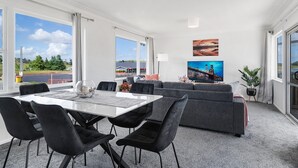 Uma sala de estar moderna com um sofá cinzento, uma mesa de jantar com cadeiras pretas e uma janela ampla com vista para o exterior.