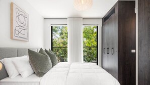 Ein Schlafzimmer mit einem großen Bett, einem grauen Kopfteil, einer weißen Wand mit einem gerahmten abstrakten Kunstwerk, einem Fenster mit Blick auf Bäume und einem dunklen Kleiderschrank.