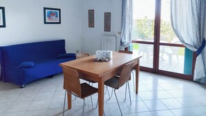 Una habitación con sofá azul, mesa de comedor de madera, sillas de mimbre y un ventanal con cortinas.