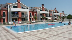Zona de piscina con suelo de baldosas, una alberca y un edificio con balcones y mobiliario exterior.
