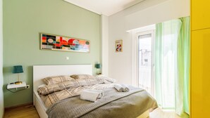 Un dormitorio con cama, mesita de noche, lámpara, un cuadro y una ventana con cortinas.