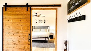 Una habitación con cama, ventilador de techo, cartel de bienvenida y perchero en la pared.