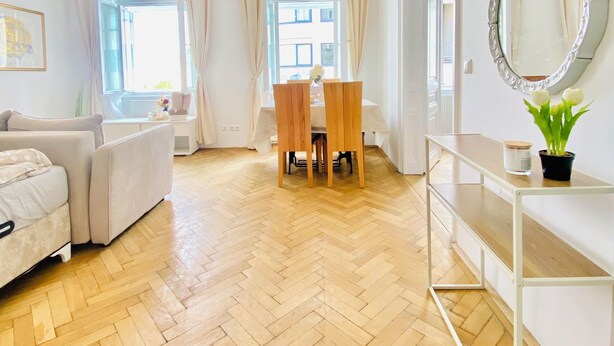 A modern living room with a herringbone wooden floor, a white sofa, a dining table with chairs, a pendant light, and a mirror on a console table. - Affordable 2BR Apt For 6 Near Stadthalle (Vienna)