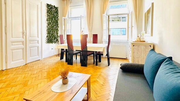 A spacious living area with a wooden herringbone floor, a sofa, a dining table with chairs, and a view of the outside through large windows. - Comfortable 1BR Lounge Westbahnhof (Vienna)