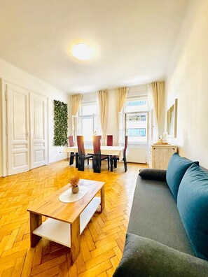 Een ruime woonkamer met een houten visgraatvloer, een bank, een eettafel met stoelen en een uitzicht buiten door grote ramen.