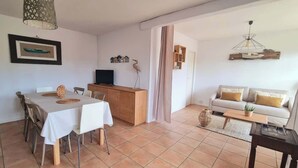 A living room with a sofa, a dining table with chairs, a TV, and a kitchen area with a cabinet and a decorative bird statue. - Charming Arès Villa: Beach, BBQ, & Fireplace! (Arès)