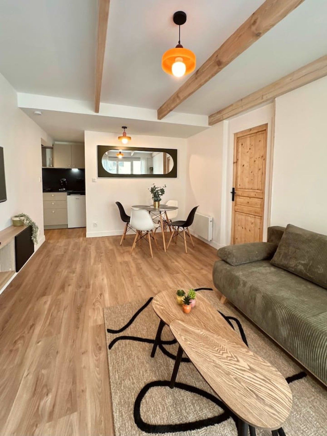 Un salon moderne avec un plancher en bois, un canapé vert, une table basse et un coin repas agrémenté d’un miroir et de chaises.
