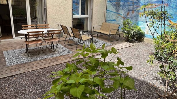 Une terrasse extérieure avec une table et des chaises, un petit jardin et un bâtiment doté d’un mur à texture et d’une fresque murale.